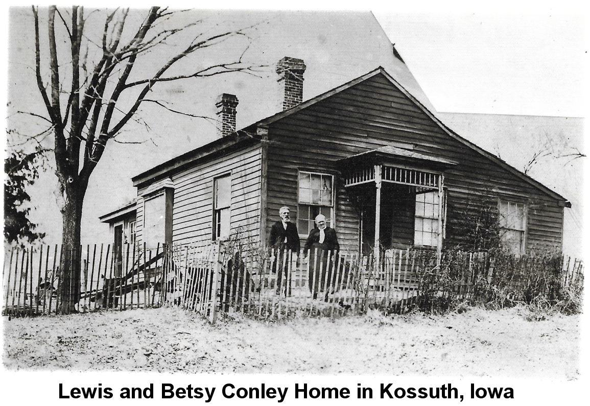 IMAGE/PHOTO: Lewis & Betsy Conley Home in Kossuth, Iowa: Black and white photo of an unpainted, rough-slab-sided one-story house with a combination peaked and shed roof and two brick chimneys, featuring an ornate but small porch roof at the front and a bay window on the left side, behind a rough picket fence, A large leafless tree stans to the left, and an elderly man with a beard stands next to an elderly woman with white hair on the front porch.